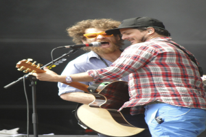 Zwei Männer auf der Bühne mit Gitarren und Mikrofonen, einer in einem rot-blau karierten Hemd und blauen Hosen, der andere mit einer Sonnenbrille und einer schwarzen Mütze.