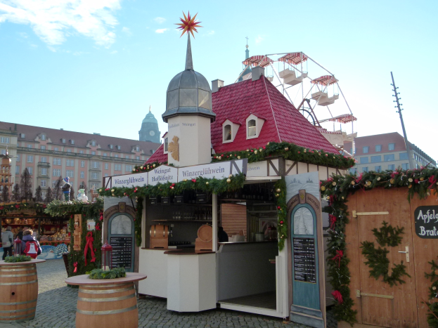 Kleines rotes Dachgebäude mit einem Riesenrad davor, umgeben von festlichen Oktoberfest-Dekorationen, Menschen und Gegenständen, vor einer Kulisse aus Gebäuden, Bäumen und einem bewölkten Himmel.