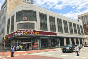 Eine belebte Stadtstraße mit Fahrzeugen und Fußgängern, hohen Glasgebäuden und einem Schild mit der Aufschrift "The Best Value Urban Outlet" unter einem bewölkten Himmel.