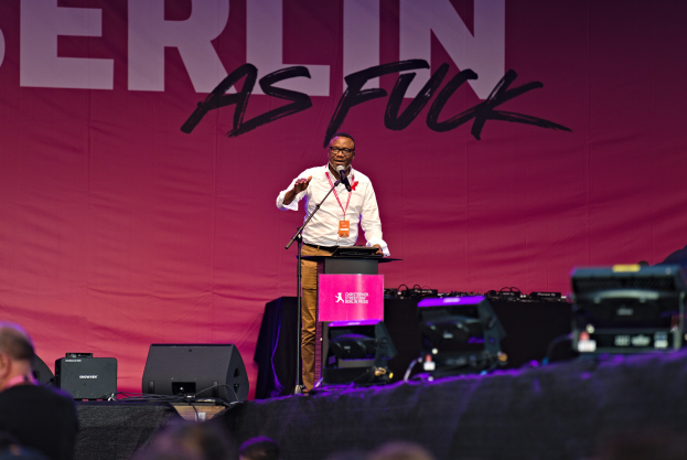 Ein Mann in einem weißen Hemd und braunen Hosen steht auf einer Bühne und hält ein Mikrofon in der Hand, während er eine Menge anspricht. Im Hintergrund ist ein pinkes Banner mit der Aufschrift 'Berlin as Fuck' zu sehen.