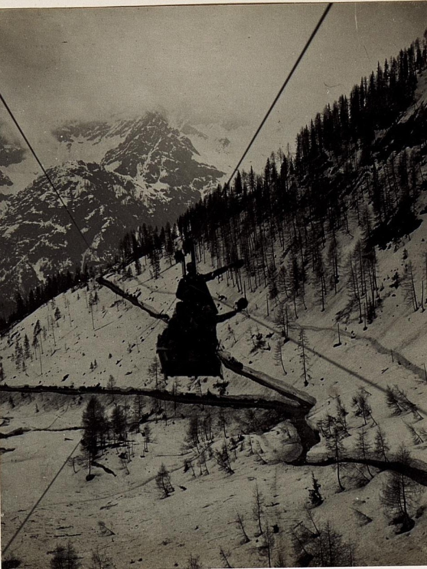 Schwarze-Weiß-Foto einer Person auf einem Skilift mit schneebedeckten Bergen und Bäumen im Hintergrund, mit Text unten.