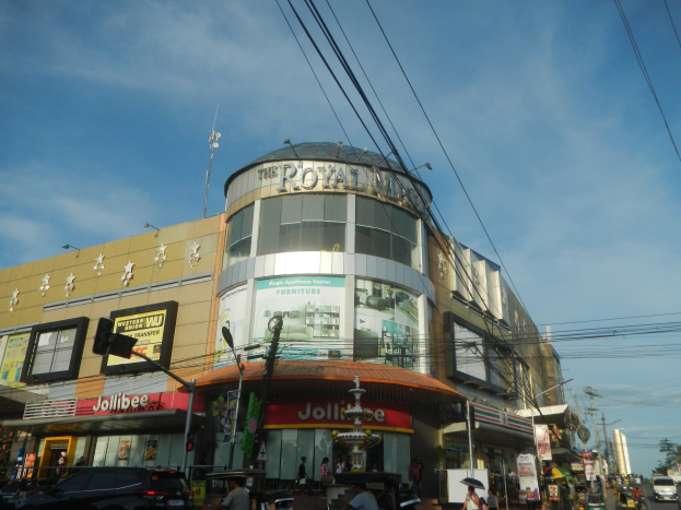 Eine belebte Stadtstraße mit fahrenden Autos, Fußgängern auf dem Gehweg und einem großen Gebäude mit der Aufschrift "Royal Mall" im Hintergrund, umgeben von parkenden Autos, Bäumen, Strommasten und Drähten unter einem klaren Himmel.