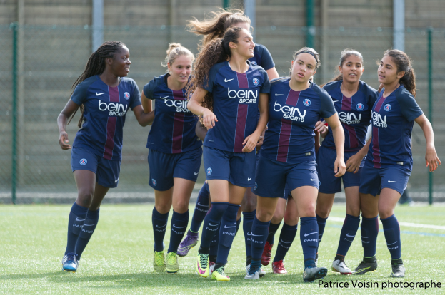 Eine Gruppe junger Frauen beim Fußballspielen auf einem Rasenfeld mit Maschendrahtzaun und einer Wand im Hintergrund, beschriftet mit "Paris Saint-Germain Frauenfußball" in der rechten unteren Ecke.