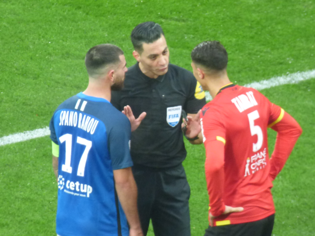 Schiedsrichter spricht mit zwei Männern auf einem Fußballfeld, einer in schwarzem Hemd und Hose, der andere in einem blauen und roten T-Shirt mit Text und Brille.