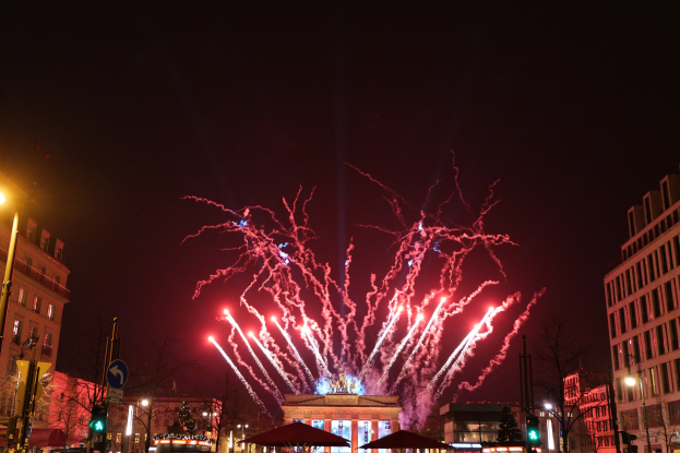 Eine belebte Stadtstraße an einem Neujahrsabend in Berlin, voller Menschen, Fahrzeuge und erleuchtet von Gebäudelichtern und Feuerwerk am Himmel.