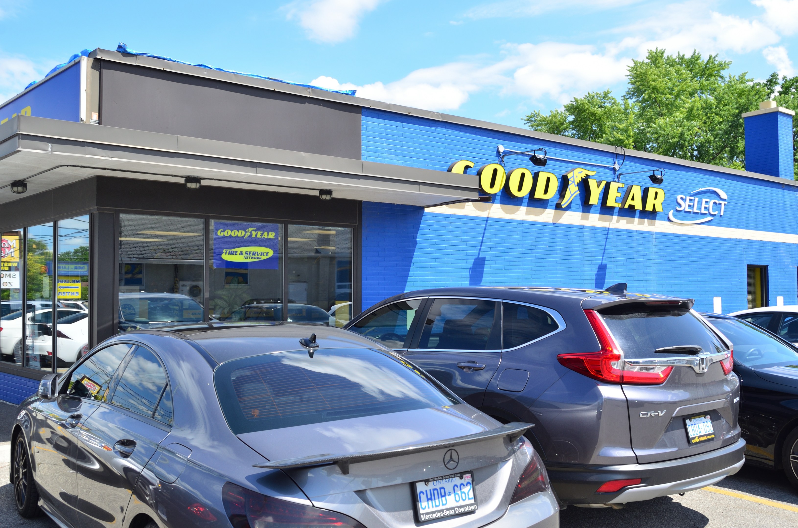 Außensicht einer Goodyear Select-Autohändlung mit parkenden Fahrzeugen davor, Beschriftung auf dem Gebäude, Bäumen im Hintergrund und einer bewölkten Himmel.