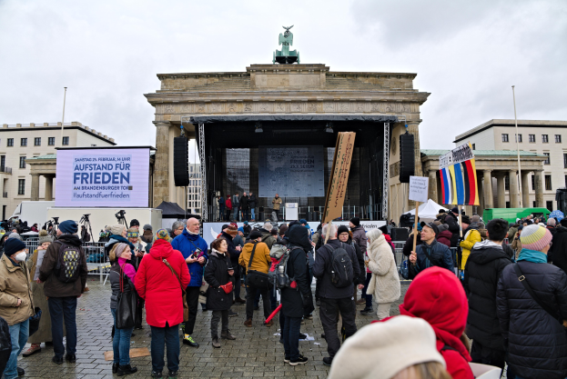 Eine Menschenmenge steht vor einem Gebäude mit einer Bühne, auf der Redner und ein Bildschirm zu sehen sind, begleitet von Fahnen und Transparenten mit Text auf der rechten Seite während einer Demonstration in Berlin.