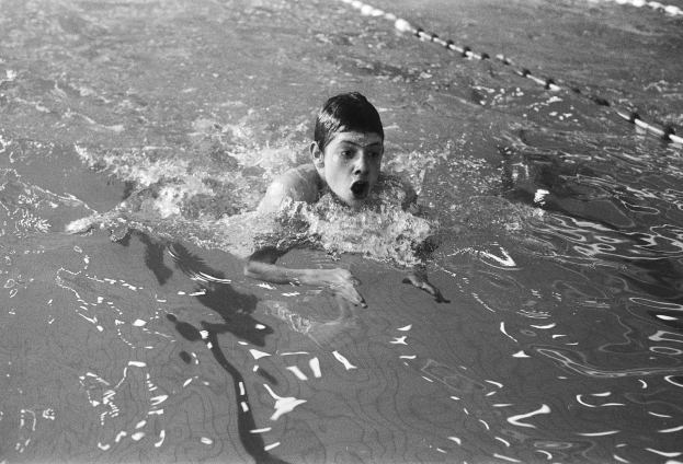 Ein junger Junge schwimmt in einem Pool mit offenem Mund, umgeben von Schwimmbahnen, in Schwarz-Weiß.