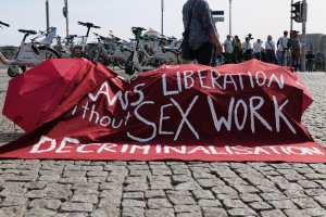 Gruppe von Menschen mit Schirmen in der Nähe von Fahrrädern mit einem roten Protestbanner, auf dem "Befreiung ohne Entkriminalisierung der Sexarbeit" steht, vor Gebäuden und Bäumen.