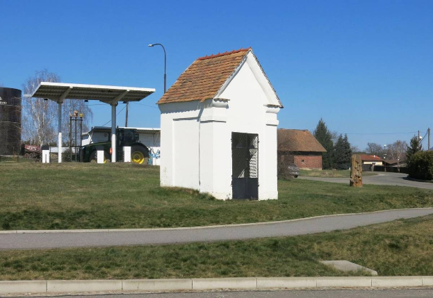 Kleines weißes Gebäude mit rotem Dach, erste Tankstelle Deutschlands, an einer grasbewachsenen Straße mit Bäumen, Strommasten und Häusern unter einem klaren blauen Himmel.