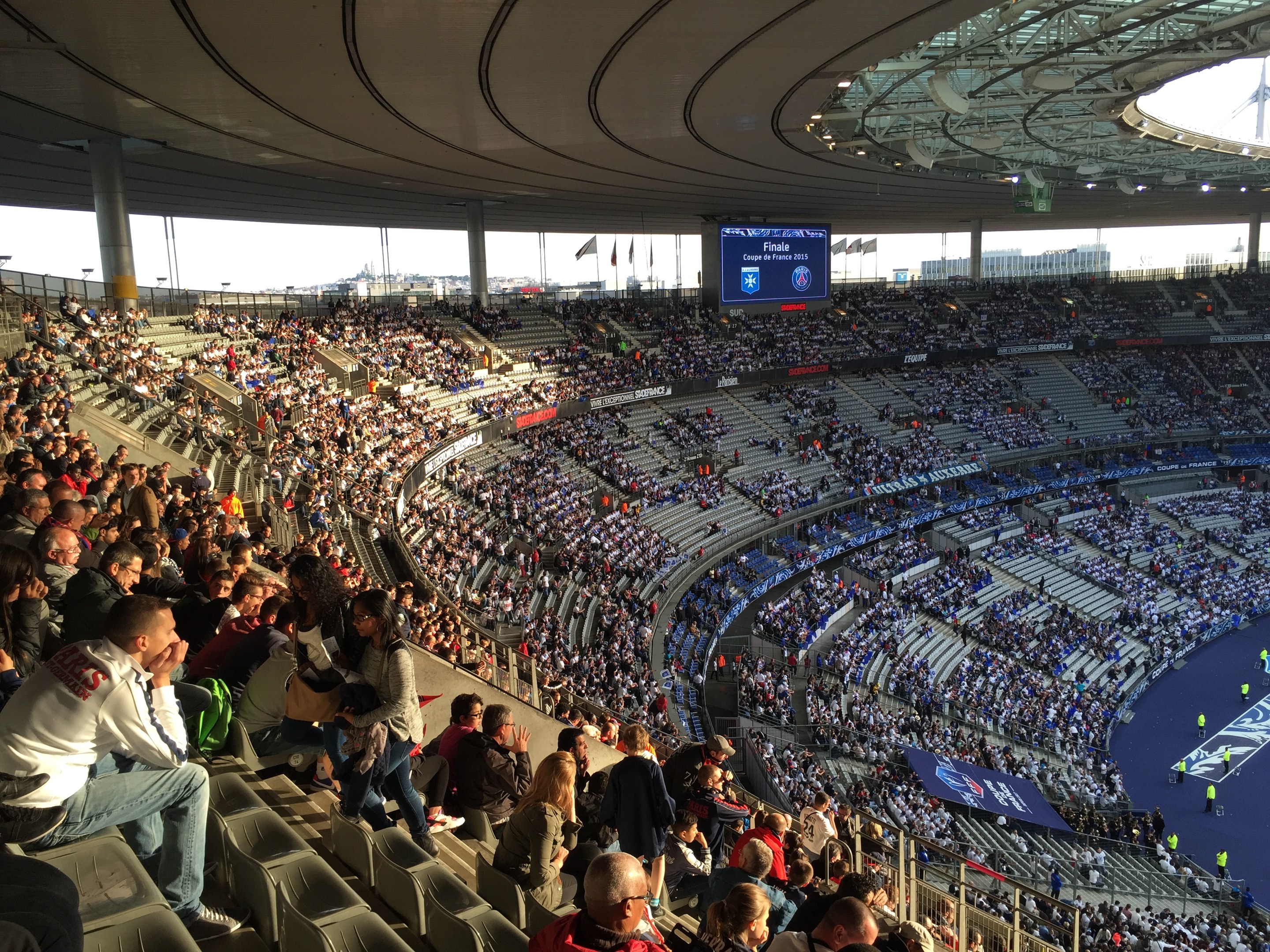 Große Menschenmenge in einem Stadion bei einem Förspiel mit einer Bühne, Fahnen, Masten, einem Bildschirm und der Allianz Arena in München im Hintergrund.