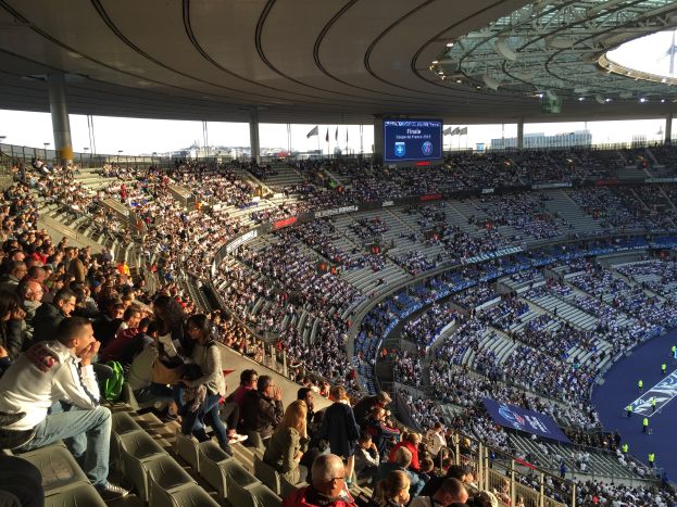 Große Menschenmenge in einem Stadion bei einem Förspiel mit einer Bühne, Fahnen, Masten, einem Bildschirm und der Allianz Arena in München im Hintergrund.