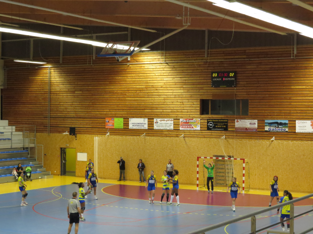 Eine Gruppe von Menschen, die Handball in einer Turnhalle mit einem Metallzaun, einem Tor, einer Tür, Texttafeln, einem Bildschirm, einer Lautsprecherbox, Metallstangen und einer Decke mit Deckenleuchten und Metallrahmen spielen.
