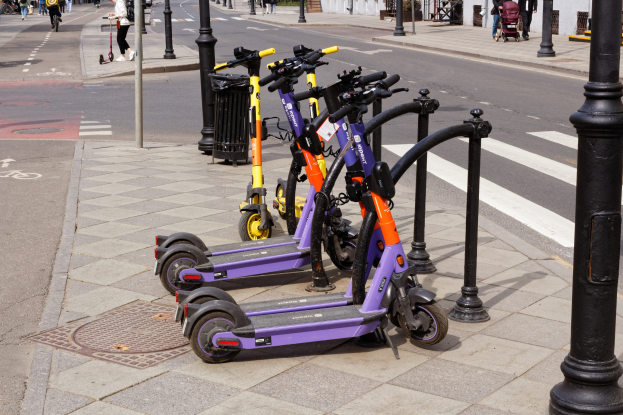 Eine Reihe von Elektro-Scootern, die an einer Straße mit Fußgängern, Radfahrern, einem Müllcontainer im Vordergrund und Gebäuden, Bäumen und Pflanzen im Hintergrund geparkt sind.