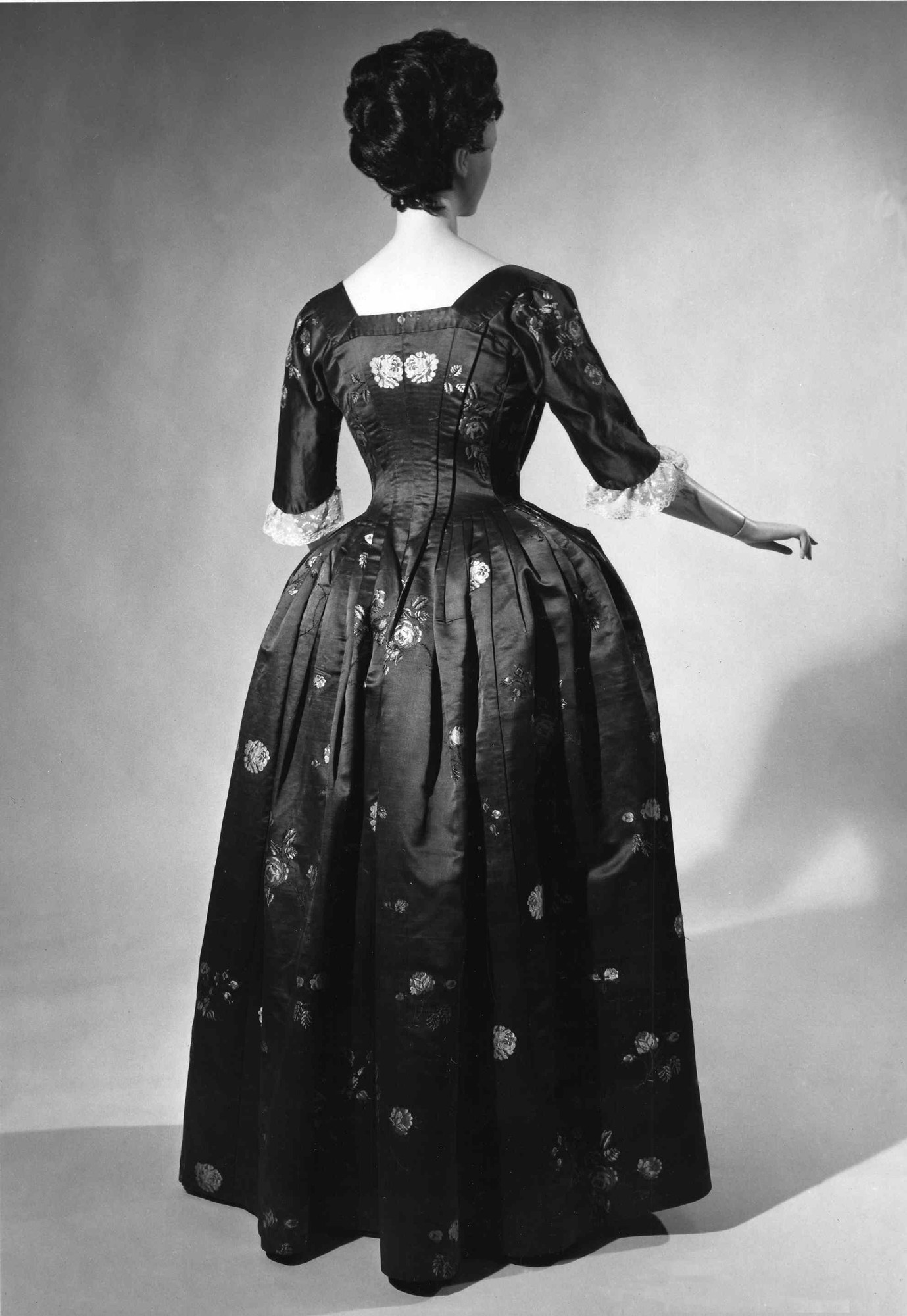 Eine Frau in einem Schwarz-Weiß-Foto aus den 1950er Jahren trägt ein enges, bodenlanges Kleid mit luxuriösem Stoff, verschränkten Händen, einem Hochsteckdutt, schwarzen Pumps und dezentem Make-up.