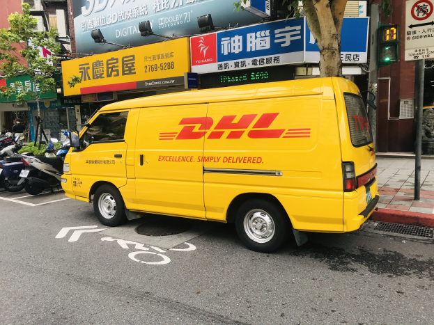 Ein gelber DHL-Lieferwagen parkt auf einer Straße neben Gebäuden, Bäumen, Schildern, Polen, Ampeln und einem Fußgänger.