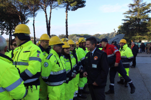 Eine Gruppe von Menschen in gelben Westen und Helmen, einige mit Mützen, steht zusammen auf einer Straße umgeben von Bäumen unter einem klaren blauen Himmel.