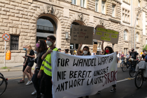 Eine Gruppe von Menschen marschiert auf einer Demonstration durch die Straße, einige halten Schilder und andere fahren Fahrräder, mit einem Gebäude mit Fenstern, Bögen, Säulen und Skulpturen, Bäumen und einer Straßenlaterne im Hintergrund.
