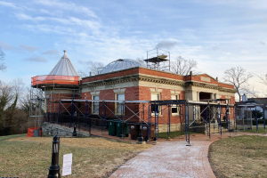 Ein großes Backsteingebäude mit Kuppel, umgeben von einem Metallzaun, einem Weg, Gras, einem Hinweisschild, Mülltonnen, Bäumen und Häusern unter einem bewölkten Himmel, scheint restauriert zu werden.