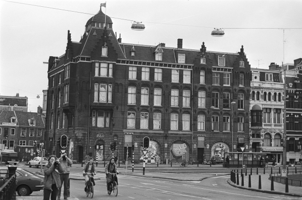 Schwarz-weiß-Foto von Menschen, die Fahrräder auf einer Stadtstraße fahren, mit Gebäuden, Straßeninfrastruktur, Kraftfahrzeugen, Fußgängern und Himmel.