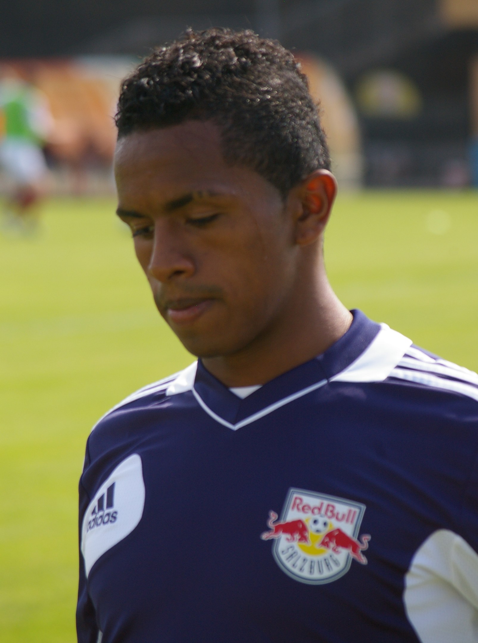 Junger Mann in blauem T-Shirt auf einem Fußballfeld stehend, der als Flügelspieler mit kurzen dunklen Haaren und entschlossener Miene erscheint.