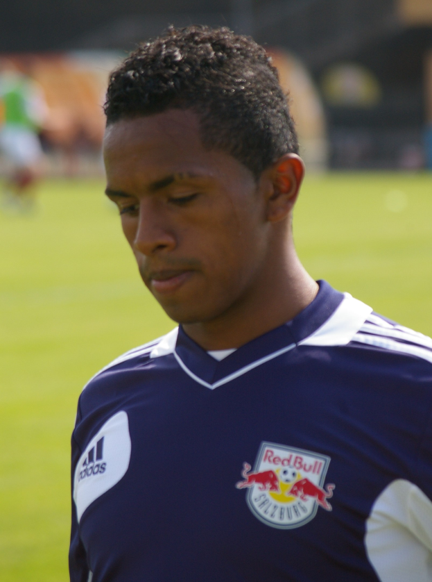 Junger Mann in blauem T-Shirt auf einem Fußballfeld stehend, der als Flügelspieler mit kurzen dunklen Haaren und entschlossener Miene erscheint.