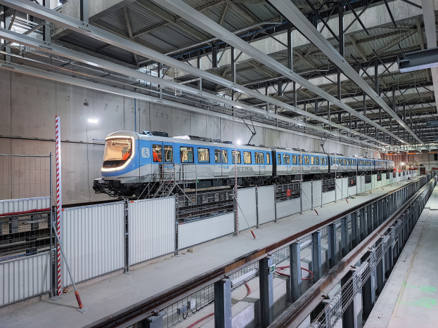 Blauer und weißer Zug fährt durch eine große Bahnhofshalle mit einer Plattform auf der rechten Seite, einer Wand im Hintergrund und einer Decke mit Stangen und Lampen.