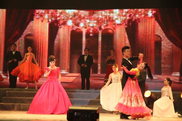 Eine Gruppe von Menschen steht auf einer Bühne mit Stufen und einem Hintergrundbild, beleuchtet von Scheinwerfern, mit einem Lautsprecher unten, was eine Tanzvorstellung suggeriert.