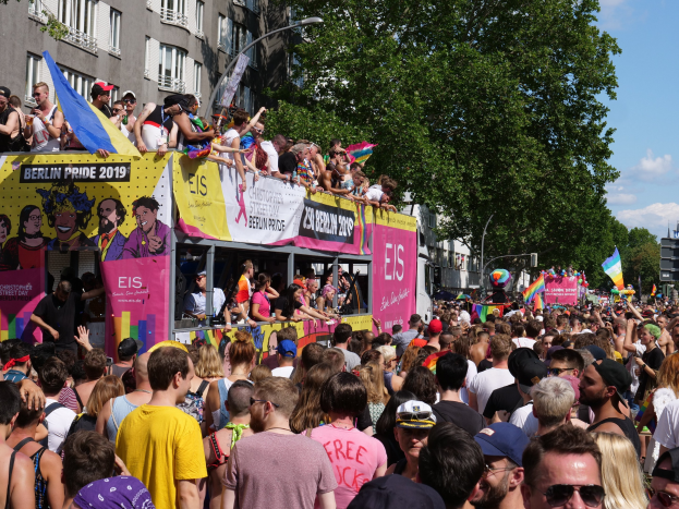 Eine große Menge marschiert während des Berlin Pride 2019 die Straße entlang, viele tragen Kappen und Schutzbrillen, einige halten Fahnen, mit Bannern, Laternenpfählen, Bäumen und Gebäuden im Hintergrund bei einem bewölkten Himmel.