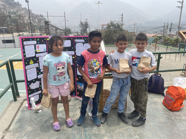 Gruppe von Kindern auf einem Basketballfeld mit Papiertüten in den H├Ąnden, mit Tüten und Gegenst├Ąnden auf der rechten Seite und einer Tafel mit Text auf der linken Seite, vor dem Hintergrund von Z├Ąunung, Masten, Stra├čenlaternen, B├Ąumen, Geb├Ąuden, H├Ąngen und Himmel.