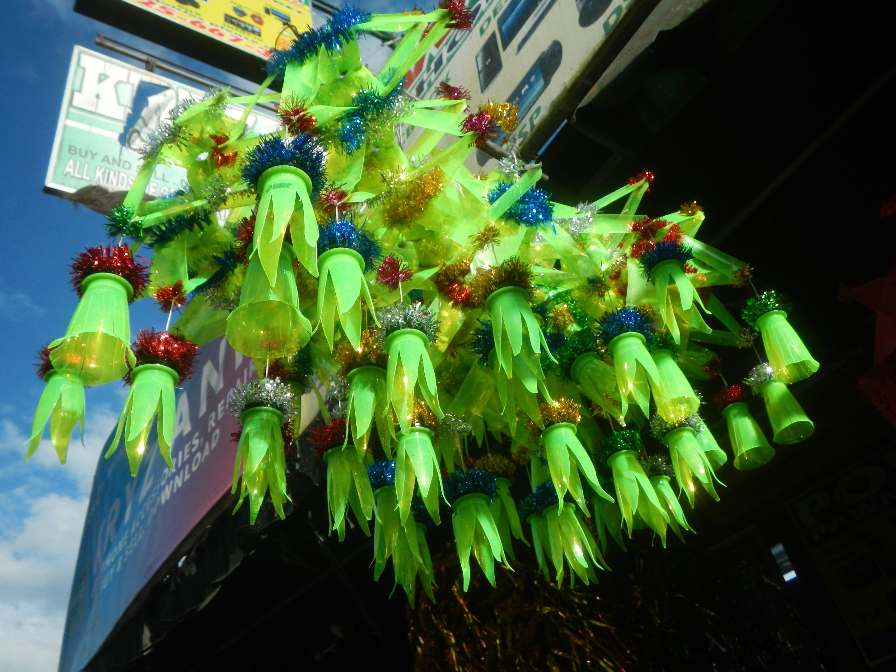 Ein festlicher Weihnachtsbaum aus grünen Plastikflaschen hängt an der Seite eines Gebäudes vor einem klaren blauen Himmel, mit einem Werbeplakat im Hintergrund.