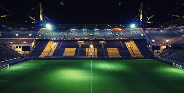 Ein hell erleuchtetes Fußballstadion bei Nacht mit saftig grünem Rasen und einigen Zuschauern auf den Tribünen.