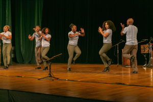 Eine Gruppe von Menschen auf einer Bühne mit Mikrofonen und Musikinstrumenten, die klatschen und feiern, mit einem Vorhang im Hintergrund.