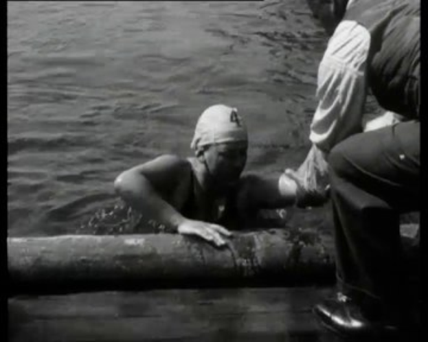 Ein Schwarz-Weiß-Bild eines Mannes in Not im Wasser mit einem Verband am Kopf, der von zwei Personen auf der rechten Seite unterstützt wird.