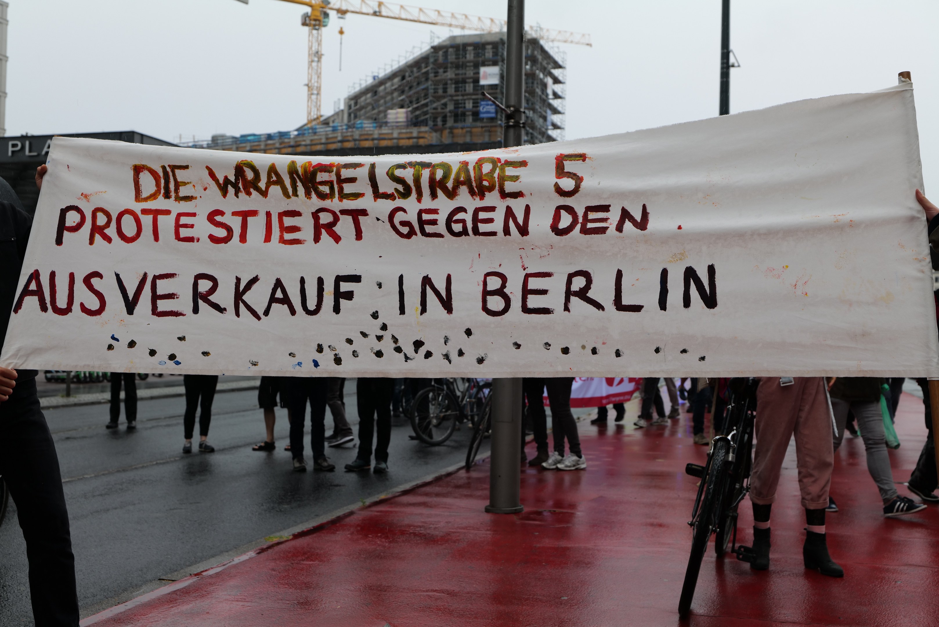 Gruppe von Menschen mit einem Banner "Die Wrangellstraße 5 Protestiert Gegen den Ausverkauf in Berlin" auf einer Straße mit Fahrrädern, Pfählen, Gebäuden und einem Kran im Hintergrund.