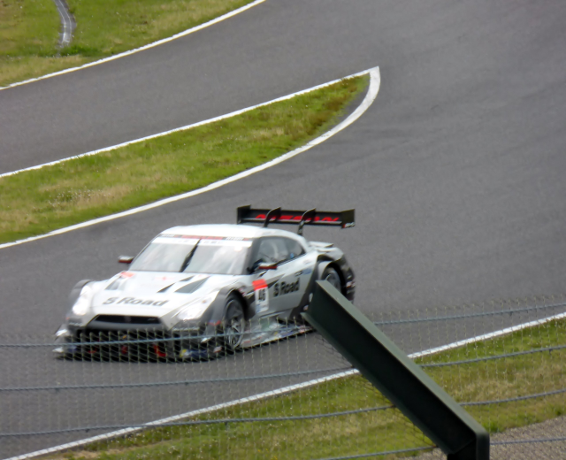 Audi R8 LMS GT3 Rennauto auf der Nürburgring 24 Stunden Strecke positioniert, umgeben von einem Metallzaun und grünem Gras, frontal auf der Asphaltstrecke ausgerichtet.