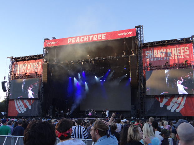Eine große Menschenmenge steht vor einer hell erleuchteten Bühne auf dem Shaky Knees Music Festival, auf der Musiker unter Textbannern spielen, mit einem Geländer im Vordergrund und einem klaren blauen Himmel im Hintergrund.