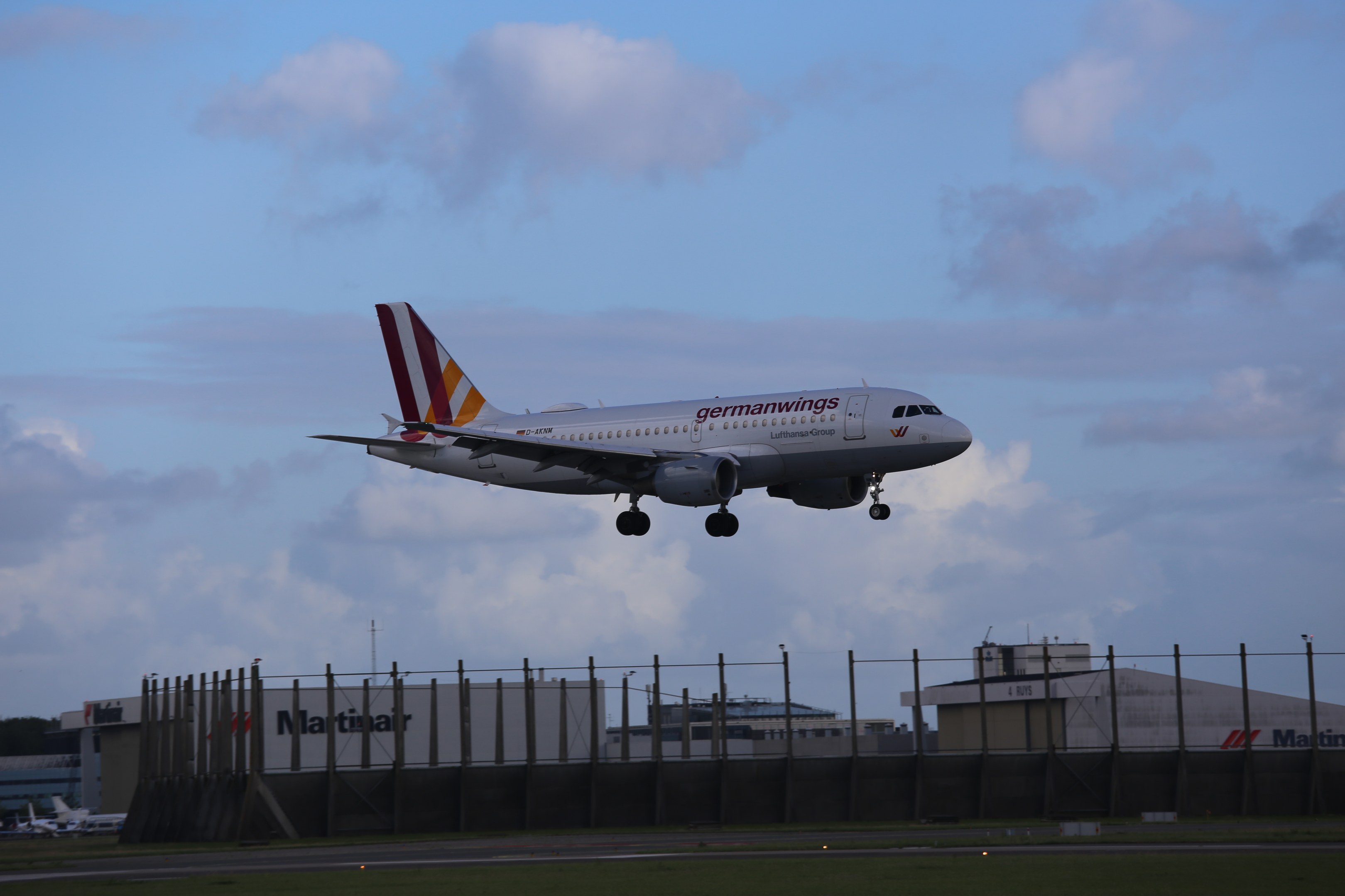 Ein Airbus A320-200 der Germanwings beim Abheben vom Frankfurter Flughafen, mit Gras, einem Zaun, Gebäuden, Bäumen und einem bewölkten Himmel im Hintergrund.