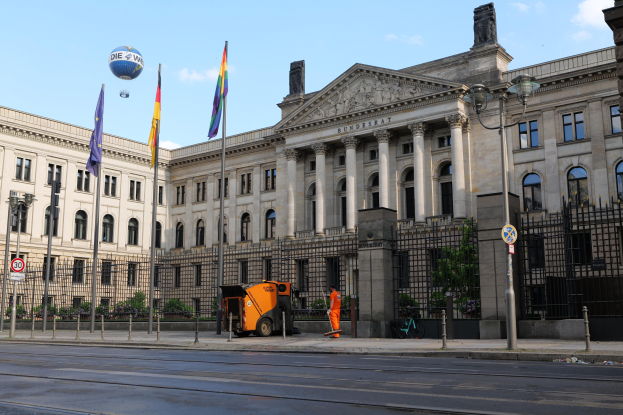 Großes modernes Regierungsgebäude mit vielen Fenstern, umgeben von Straßeninfrastruktur und Fußgängern, vor einem bewölkten Himmel.
