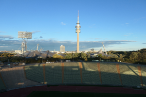 Olympiastadion in Berlin mit dem Fernsehturm (Fernsehturm) im Hintergrund, umgeben von Bäumen, Gebäuden und beleuchteten Bereichen unter einem bewölktem Himmel.
