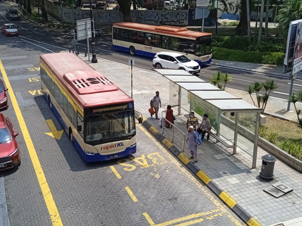 Stadtstraße mit fahrenden Autos, Bussen, einer Bushaltestelle, Fußgängern auf dem Gehweg, Strommasten, Schildern, Bäumen, Gras und umliegenden Gebäuden.