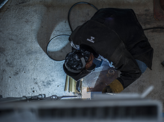 Ein Schweißer in Schutzausrüstung bedient eine Schweißmaschine an einem Metallstück, mit Werkzeugen und Gegenständen, die auf dem Boden verstreut sind.