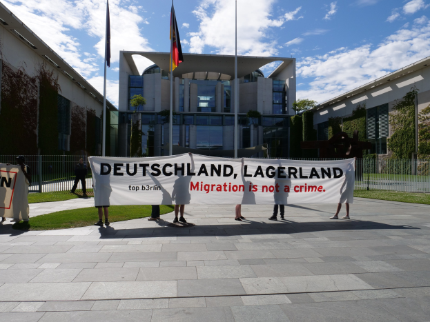 Gruppe von Menschen mit einem Banner mit der Aufschrift 'Deutschland, Lagerland Migration ist kein Verbrechen' vor einem Gebäude mit Fenstern, umgeben von Gras, einem Metallzaun, Pflanzen, Bäumen und Fahnen auf Stangen unter einem bewölkten Himmel.